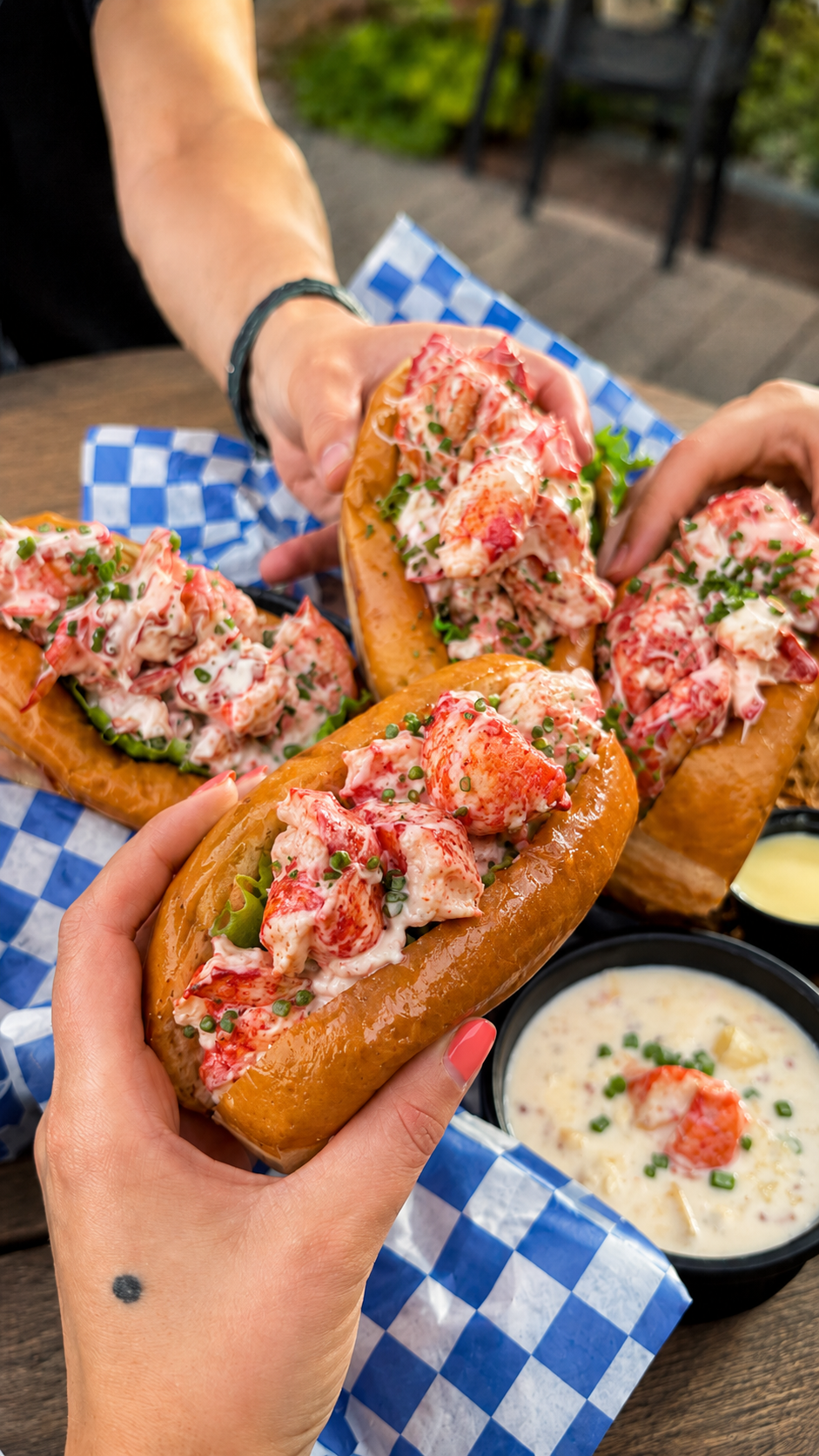 Friends toasting and holding lobster rolls together over an outdoor picnic table spread with clam chowder